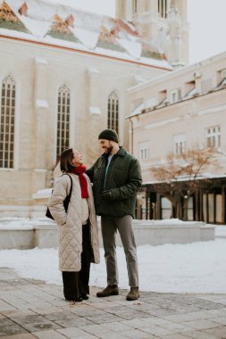 Karlı bir günde bej ceket ve kırmızı eşarp giyen Koreli bir kadın yeşil ceketli beyaz bir adamla duruyor. Avrupa 'da bir kilisenin önündeler..