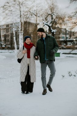 Gülümseyen Koreli bir kadın ve beyaz bir adam karlı bir parkta el ele yürüyorlar. Kadın uzun bir ceket ve kırmızı eşarp giyiyor. Soğuk bir kış günü gibi görünüyor..