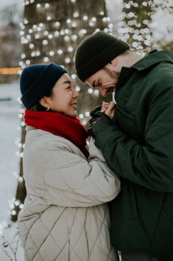 Koreli bir kadın ve beyaz bir erkek birbirlerine yakın dururken birbirlerine gülümsüyorlar. Sıcak giyimli ve etrafı ışıklı ağaçlarla çevrili. Kış geldi ve mutlu görünüyorlar..