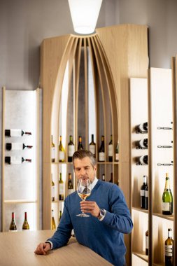 Ahşap bir barda elinde şarap kadehi tutan bir adam var. Onun arkasında çeşitli şarap şişeleriyle dolu raflar var. Ayar parlak ve bar alanına odaklı..