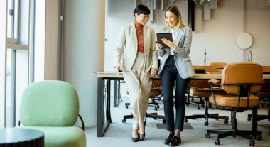 Çağdaş bir ofiste iki iş kadını birlikte tablete bakarken gülümsüyor. Etraflarında gösterişli, şık bir çalışma alanı, toplantı sandalyeleri ve masaları var..