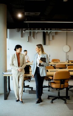 İki kadın bir ofiste yürüyor ve konuşuyor. İş kıyafetleri giymişler. Arka planda masalar ve sandalyeler var ve diğer insanlar çalışıyor..