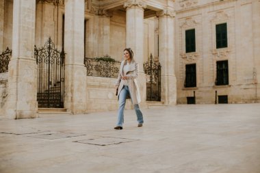 Malta 'da taş bir sokakta yürüyen bir kadın. Geniş kolonları ve detaylı kapıları olan eski binaların yanından geçiyor. Dışarıda geçirdiği zamandan zevk alırken güneş onun üzerine doğuyor..