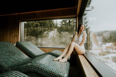 Bir kadın, kaplıca alanında bir çıkıntıda oturur ve dışarıdaki karlı ağaçların tadını çıkarır. Basit bir kıyafet giymiş ve pencere pervazında otururken rahatlamış görünüyor..