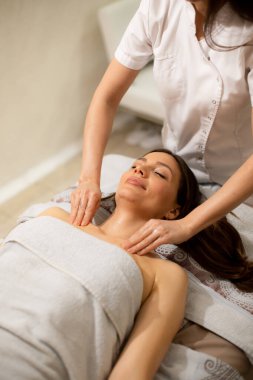 Bir kadın masaj masasında yatarken bir başkası ona masaj yapıyor. Ortam bir sağlık merkezi. Atmosfer rahatlama ve bakıma odaklı..