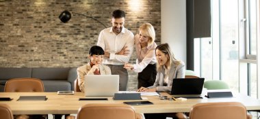 Bir grup iş arkadaşı bir ofisteki masanın etrafında toplanıyor. Fikirlerini paylaşıyorlar ve bilgisayarları ve kağıtları kullanarak işleri tartışıyorlar. Ayar parlak ve modern bir tasarıma sahip..