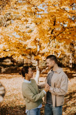 Koreli bir kadın ve beyaz bir erkek, sakin bir parkta altın sonbahar yaprakları arasında bir an için gülüyorlar..