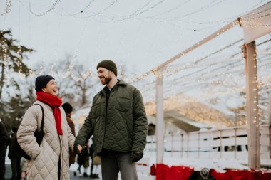 Koreli bir kadın beyaz bir adamla konuşurken gülümsüyor. Işıklarla dolu bir açık hava etkinliğindeler. Dışarısı soğuktayken insanlar kalın giysiler içinde dolaşır..