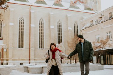 Koreli bir kadın ve beyaz bir adam Avrupa 'da bir kilisenin yanındaki karlı bir sokakta el ele yürüyorlar. İkisi de gülümsüyor ve kalın kışlıklar giyiyorlar..