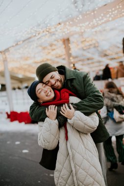 Koreli bir kadın ve beyaz bir erkek kış pazarında kucaklaşırken birlikte gülümsüyorlar. Sahne tatil ışıkları, insanlar ve dekorasyonlarla dolu..