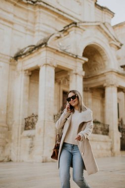 Malta 'da bir kadın telefonla konuşurken sokakta yürüyor. Klasik mimarisi olan eski bir binanın yanında. Gündüz oldu ve güneş parlıyor..