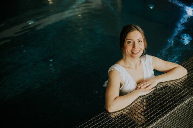 Bir kadın havuzun kenarına yaslanırken gülümser. Su onun etrafındaki ışığı yansıtıyor. O, basit bir kıyafet giydi ve rahat görünüyor. Bu sahne kapalı bir yerde geçiyor..