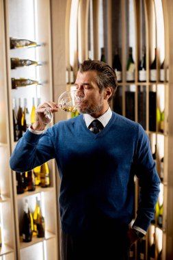 Bir adam şık bir şarap mahzeninde duruyor. Dudaklarına bir bardak beyaz şarap dayayıp tadını çıkartıyor. Mahzende arka planda yığılı bir sürü şişe var..