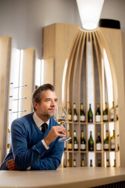 Bir adam elinde bir bardak şarapla bir barda duruyor. O, cama bakarken düşünceli görünüyor. Arkasında şarap şişeleri var. Dostça bir atmosfer yaratıyor..