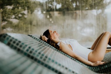 Bir kadın spa 'da şezlongda yatıyor. O bir mayo giyiyor ve gülümseyerek yukarı bakıyor. Arkasından su akıyor, rahatlatıcı bir atmosfer yaratıyor..