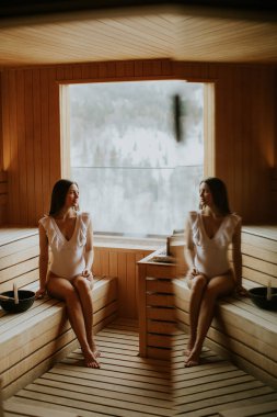 Bir insan sıcak tonlar ve yumuşak ışıkla çevrili ahşap bir saunada oturur. Pencereden görünen manzara dışarıdaki karlı manzarayı gösteriyor. Sakin alan, yansımayı ve rahatlamayı davet eder..
