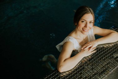 Bir kadın kapalı havuzda rahatlar. Usulca gülümseyerek kenarlara yaslanıyor. Su yumuşak ışığı yansıtır. Bu sahne, akşam vakti özel bir ortamda neşeli bir an gösteriyor..