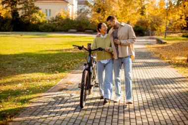 Koreli bir kadın ve Kafkasyalı bir erkek, sonbahar parkında düşmekte olan yapraklarla çevrili bir bisikletin yanında yürürken gülüyorlar..