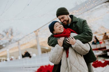 Koreli bir kadın ve beyaz bir erkek açık hava pazarında sıcak bir anı paylaşıyorlar. Neşeli ışıklar bölgeyi süslerken gülümsüyor ve kucaklaşıyorlar. Çevrelerinde, kış süslemeleri vardır..