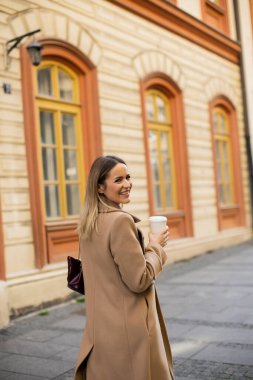 Bir kadın kaldırım taşı caddesinde yürürken gülümsüyor. O bir kahve fincanı tutar ve bir ceket giyer. Etrafındaki binaların parlak renkleri ve geniş pencereleri var..