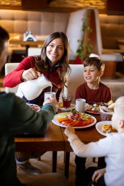 Bir kadın oğlu için süt dökerken genç bir çocuk kafede izliyor. Masa yiyecek ve içeceklerle dolu. Hepsi gülümsüyor ve birlikte sabahtan zevk alıyorlar..
