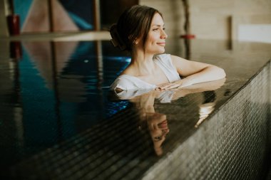 Bir kadın sakin suyla dolu bir havuzda vakit geçirmekten hoşlanır. Düşünceli bir şekilde bakarken kenara yaslanıyor. Ayar parlak ve temiz çizgileri var. Rahat bir atmosfer yaratıyor..