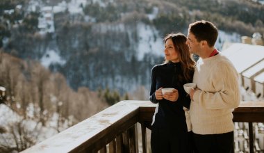 Bir çift birlikte balkonda durur, ılık içecekler yudumlarlar ve sakin bir kış gününde karlı dağ manzarasının tadını çıkarırlar..