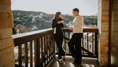 İki kişi bir balkonda durmuş, kahve içip sohbet ediyorlar. Güneş arkadaki karlı dağların üzerinde parlıyor. Kış boyunca aydınlık ve soğuk bir sabah..