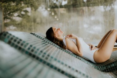 Bir kadın kaplıcada sıcak taştan bir yatakta yatıyor. Mayo giyiyor ve dinlenirken gülümsüyor.