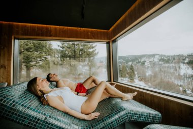 İki kişi sıcak taştan bir yatakta spa 'da uzanıyorlar. Soğuk bir kış gününde büyük pencerenin dışındaki beyaz kar ve ağaçların tadını çıkarırken rahatlamış görünüyorlar..
