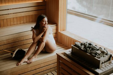 Bir kadın saunada ahşap bir bankta oturuyor, rahat ve düşünceli görünüyor. Sıcak odun ve taşlarla çevrili bir sağlık merkezinde. Pencereden güneş ışığı süzgeçleri.