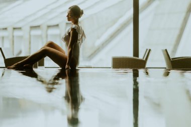 Modern bir binanın yansıtıcı yüzeyinde oturan bir kadın. Güneş ışığı bölgeyi doldururken o sessizliğin tadını çıkarıyor. Bu geniş ortamda onun etrafında sandalyeler düzenlenir..