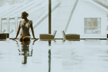 Bir kadın kapalı havuzun kenarında oturuyor. Yağmur yağarken dışarıya bakıyor. Sandalyeler yakınlarda. Ortalık sakin ve hava güzel görünüyor..