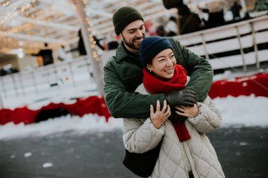 Koreli bir kadın ve beyaz bir erkek açık hava kış etkinliğinde gülümserken kucaklaşıyorlar. Etrafları süslemelerle çevrili ve insanlar festival atmosferinin tadını çıkarıyorlar..