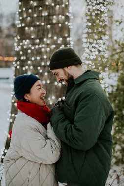 Koreli bir kadın ve beyaz bir erkek parkta birbirlerine yakın dururlar. Çevrelerindeki ağaçlar parlak ışıklarla aydınlatılmış. Gülümsüyorlar ve açık havada kış gününün tadını çıkarıyorlar..