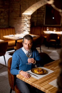 Bir adam bir restoranda ahşap bir masada tek başına oturuyor. Bir bardak su tutuyor ve yemek tabağına bakıyor. Dekor rahat bir atmosferle iyi aydınlatılmış..