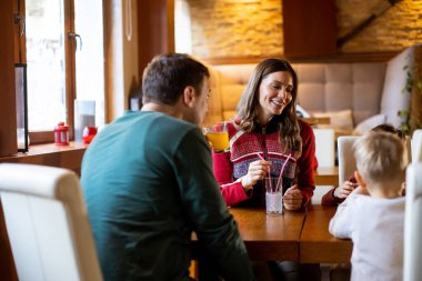 Bir aile bir restorandaki ahşap bir masada oturur. Bir kadın elinde içki tutarken gülümsüyor. Bir adam ve küçük bir çocuk anı paylaşıyorlar. Birlikte vakit geçirmekten zevk alıyor gibiler..