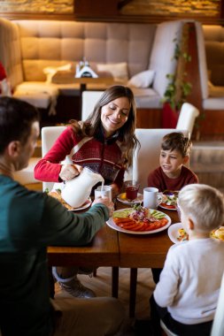 Bir aile restoranda bir masada oturur. Anne onlar kahvaltıyı paylaşırken oğluna bir içki koyar. Masada yemek tabakları var. Sabah oldu ve çok mutlular..