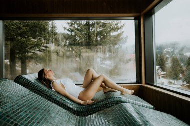 Huzurlu bir kadın, büyük pencerelerden nefes kesici bir dağ manzarası seyrederken, kaplıcadaki sıcak taş bir yatağa uzanıp sükuneti kucaklıyor..