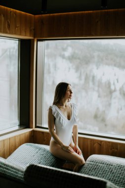 Sıcak bir taş yatakta huzur içinde oturan bir kadın rahatlatıcı bir spa 'da, karlı dağları büyük pencerelerden seyrediyor..