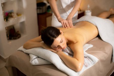 Terapist sırtına baskı uygularken bir kadın masaj masasına uzanır. Odada hafif ışıklandırma ve sakinleştirici dekor var. Oturumda rahatlama ve gerginlikten kurtulma üzerinde duruluyor.