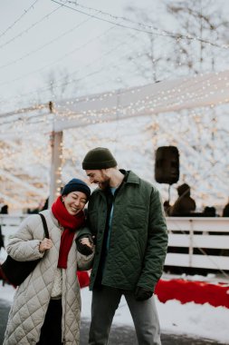 Bir Koreli kadın ve beyaz bir erkek, bir kış festivalinde el ele tutuşurken gülümsüyorlar. Kalın giysiler giyerler ve süslemelerin önünde ışıklarla dururlar. Kar zemini kaplıyor..