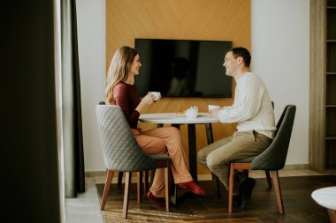 Küçük yuvarlak bir masada oturan bir çift sıcak bir mekanda kahvenin tadını çıkarıyor. Ellerinde bardak tutarken gülümser ve konuşurlar. Pencereden gelen güneş ışığı havayı canlandırıyor..