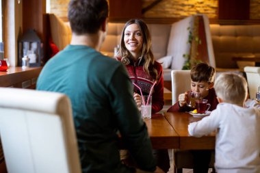Bir kadın bir adama gülümserken iki çocuk masada fincanlardan su içiyor. Yoğun bir kafede sıcak ışıklandırmayla oturuyorlar. Güzel bir öğleden sonra..