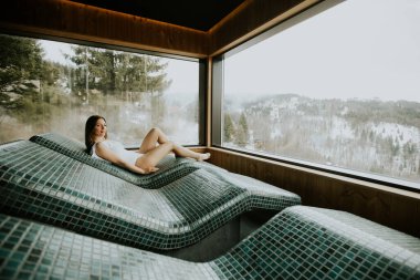 Bir kadın, sıcak taştan bir yatakta, dışarıda kış manzarası açıkken, rahatlatıcı doğa manzarasıyla çevrili, huzurlu anlardan hoşlanır..