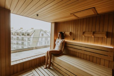 Bir kadın, ahşap bir saunada huzur içinde oturur, panoramik şehir manzarasının keyfini çıkarır, sıcaklığın tadını çıkarır..