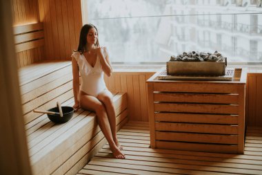 Genç bir kadın saunada sükunetin tadını çıkarıyor. Işığın büyük pencerelerden içeri akması gibi suyu yudumlayarak dışarıdaki şehri ortaya çıkarıyor..