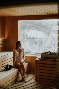 Genç bir kadın, ahşap bir saunada huzurlu bir anın tadını çıkarıyor. Büyük pencereden karlı dağlara bakıyor..