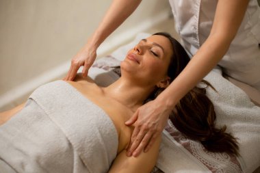 Bir kadın masaj masasına uzanmış, vücudunda bir havluyla. Bir terapist ellerini omuzlarına masaj yapmak için kullanır. Ortam sakin ve rahatlık ve rahatlığa odaklı..