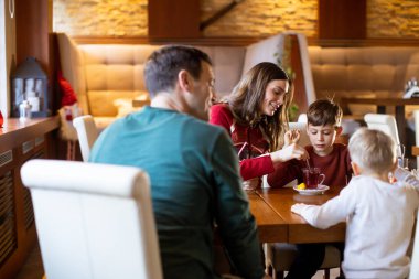 Bir aile bir restorandaki ahşap yemek masasının etrafında toplanır. Bir kadın genç bir çocuğa içkiyle yardım ederken diğer çocuk izliyor. Hava sıcak ve davetkar..